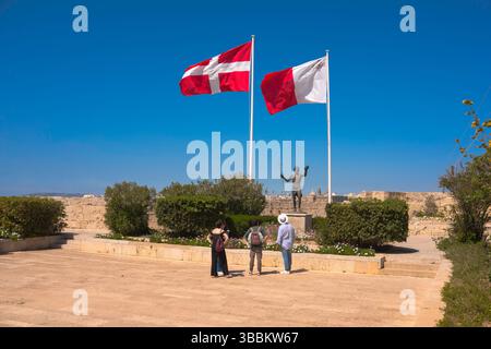 Malta, Touristen studieren eine Statue des heiligen Johannes des Täufers, flankiert von den Flaggen des Malteserordens und der maltesischen Flagge, Fort St. Angelo Stockfoto