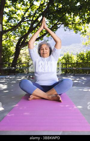 Seniorin übt Yoga mit Metallgeländer auf der Terrasse, mit rosa Yogamatte und Wasserflasche Stockfoto