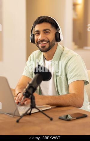 Junger erwachsener indischer Mann, der im Heimstudio aufnimmt, auf einem Laptop mit Kopfhörern und Mikrofon schreibt Stockfoto