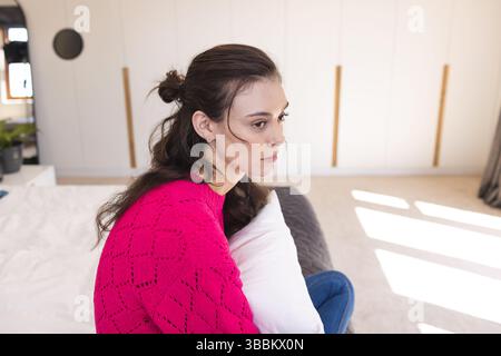Frau, die auf dem Bett sitzt und ein weißes Kissen in einem hellen, modernen Schlafzimmer hält und einen rosafarbenen Strickpullover trägt Stockfoto