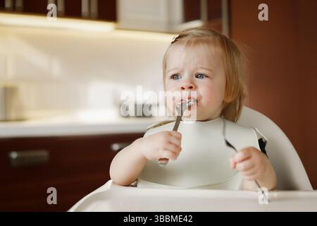 Niedliches 11-monatiges Kleinkind oder Kleinkind, das auf Hochstuhl am Fütterungstisch sitzt. Ihr Baby genießt leckeres Essen und isst selbstständig. Tochter lernen Stockfoto