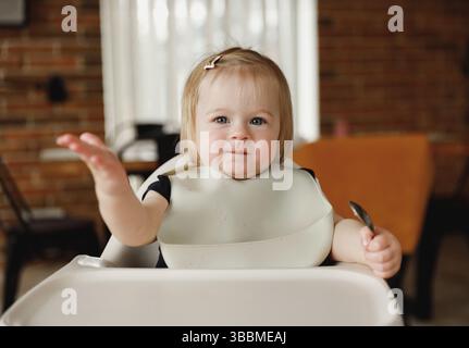 Lächelndes Kleinkind oder Kleinkind, das auf Hochstuhl am Fütterungstisch sitzt. Das Baby genießt Essen und isst selbstständig. Tochter lernt essen b Stockfoto