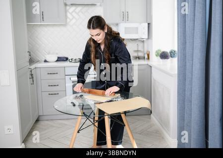 Frau, die Teig zum Backen vorbereitet, ihn zu Hause auf dem Küchentisch ausrollen. Hausfrau, die Essen zubereitet, Pasta, Pizza, Croissants, Süßigkeiten kocht. Konzeptkochen. Stockfoto