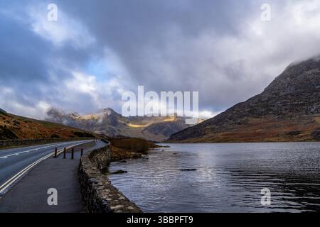 Gewundene Seestraße neben Llyn Ogwen mit gewellten Reflexen, die in Richtung der schroffen, schneebedeckten Gipfel der Glyderau unter brodelnden Wolken führen Stockfoto
