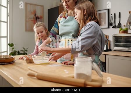Mutter und zwei Töchter backen in der Küche, messen und mischen Zutaten, zeigen familiäre Bindung und Teamarbeit, genossen Momente der Zweisamkeit, Küchenutensilien sichtbar Stockfoto