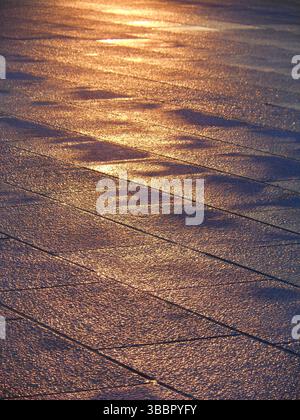 Nahaufnahme des nassen Steinpflasters, der unter Sonnenlicht glitzert, mit goldenen Highlights und unregelmäßigen Reflexen Stockfoto