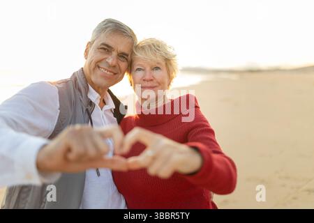 Glückliches Seniorenpaar, das bei Sonnenuntergang mit Händen am Strand Herzform macht Stockfoto