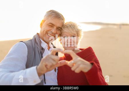Glückliches Seniorenpaar, das bei Sonnenuntergang mit Händen am Strand Herzform macht Stockfoto