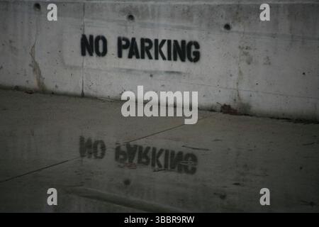 Städtische „No Parking“-Schablone, die an einem regnerischen Tag in der Stadt auf eine Betonwand gesprüht wurde, mit einer stimmungsvollen Reflexion auf dem nassen Bürgersteig. Stockfoto