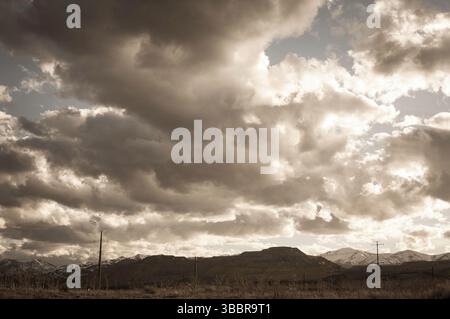 Dramatische Wolkenlandschaft über einer zerklüfteten Berglandschaft mit Versorgungsmasten am Horizont, die in Sepia-Tönen für einen stimmungsvollen und atmosphärischen Effekt gehalten werden Stockfoto