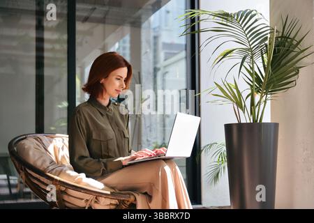 Junge rothaarige Geschäftsfrau mit Laptop, die im grünen Büro sitzt. Stockfoto
