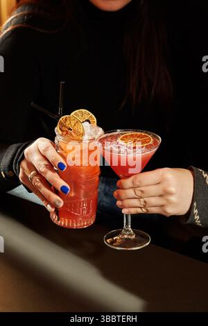 Frau, die zwei schicke Cocktails mit Orangengarnitur in einem Tiki-Becher und Martini-Glas hält Stockfoto
