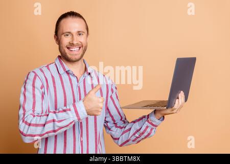 Ein lächelnder Mann im lässigen gestreiften Hemd hält den Laptop und zeigt die Daumen nach oben vor beigefarbenem Hintergrund Stockfoto