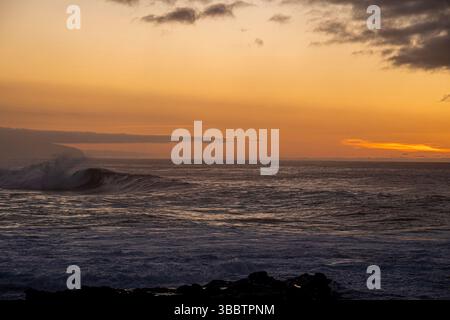Das Meer ist ruhig und der Himmel hat eine wunderschöne orange Farbe. Die Sonne untergeht und die Wellen stürzen gegen das Ufer Stockfoto