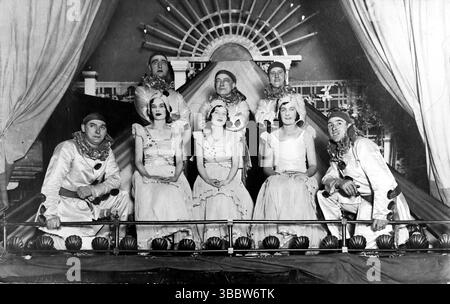 Das Bild zeigt eine Gruppe von Personen, möglicherweise Entertainern, die in einer Art Bühnenrequisite posiert, die einem großen Zelt oder Markise ähnelt. Im Vordergrund sitzt eine Frau in einem hellen Kleid, mit mehreren anderen Personen, die von hinten und über ihr hinausschauen, einige mit theatralischem Make-up oder Kostümen, darunter anscheinend Clown-Kragen auf zwei Figuren, die von den Seiten hinausgehen. Im Hintergrund befindet sich eine dekorative Struktur, die einem Ventilator oder Sonnenschliff ähnelt. Aus einer gemischten Sammlung von edwardianischen Postkarten. Stockfoto
