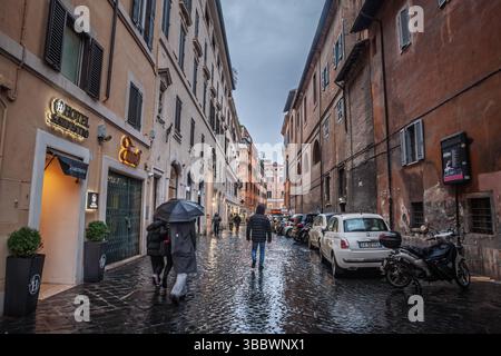 ROM, ITALIEN - 15. JANUAR 2025: Menschen, die in Mäntel mit Regenschirmen gehüllt sind, bewegen sich auf einer engen Kopfsteinpflasterstraße im historischen Zentrum Roms; feuchte Basaltsteine gleiten Stockfoto