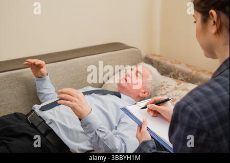 Ein älterer Mann liegt auf einer Couch und hält seine Hände während eines Termins mit einem Hypnotherapeuten hängend. Ein älterer männlicher Patient in einem Individuum Stockfoto