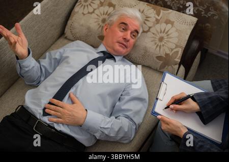 Ein älterer Mann liegt während eines Termins mit einem Psychotherapeuten auf einer Couch. Ein älterer männlicher Patient in einer individuellen Konsultation mit einer Frau Stockfoto