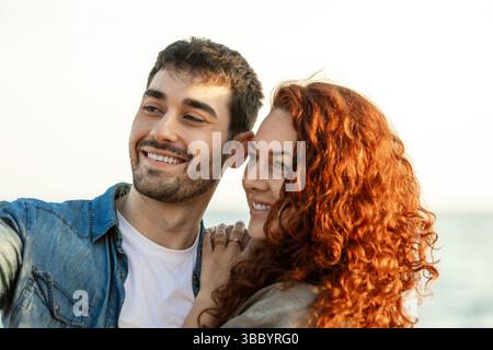 Glückliches junges Paar lächelt, während es auf das Meer blickt und sich warm umhüllt mit weichem natürlichem Licht, das ihre Gesichter und den dahinter liegenden Ozean hervorhebt Stockfoto