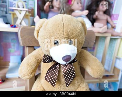Ein brauner Teddybär mit brauner Fliege sitzt auf einem Holzstuhl. Der Stuhl wird vor ein Regal gestellt, auf dem eine Puppe steht. Die Szene gibt einen ab Stockfoto