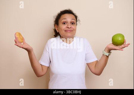 Eine Frau, die zwischen einem zuckerhaltigen Donut und einem frischen grünen Apfel streitet. Das Bild zeigt das Konzept des Gleichgewichts zwischen Genuss und gesunden Alternativen in Stockfoto