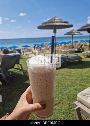 Eine Frau hält einen Cappuccino am Strand. Blauer Himmel und Regenschirme im Hintergrund. Ayia Napa, Zypern, 2023 Stockfoto