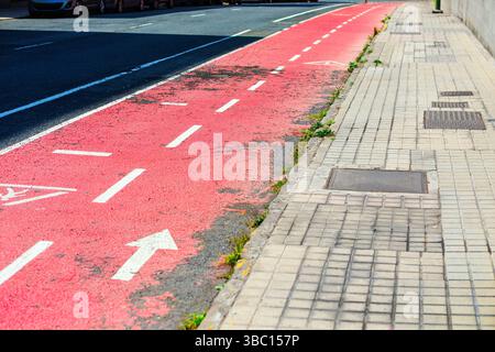 Radweg mit Anzeichen von Witterungseinflüssen, Kontrast zum grauen Bürgersteig, einer gewöhnlichen urbanen Szene. Straßenszene in der Stadt mit verblassem rosafarbenem Radweg Stockfoto