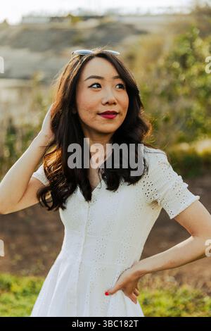 Eine asiatische Frau in einem weißen Kleid mit einem blau-weißen Muster steht auf einem Feld. Sie trägt eine Sonnenbrille und einen roten Lippenstift Stockfoto