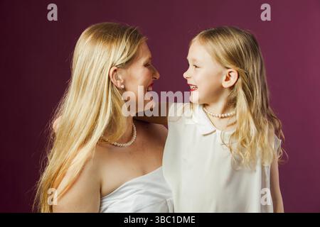 Portrait von glücklicher lächelnder Mutter und Tochter, beide mit langen, lockeren blonden Haaren, weißen Kleidern und rotem Lippenstift. Tochter sieht aus wie Mutter. St Stockfoto