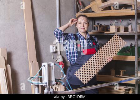 Professionelle Tischlerin in Brillen und Arbeitskleidung mit Holzgitter, die neben der Kreissäge posiert. Stereotypfreies Konzept. Zimmerlerin Stockfoto