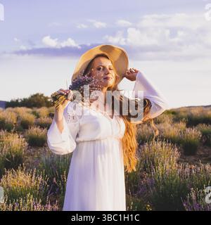 Porträt einer schönen blonden Frau, die zwischen den Plantagen von Lavendel steht. Mädchen in einem weißen Sundress gekleidet und hält sbunch Lavendel Stockfoto