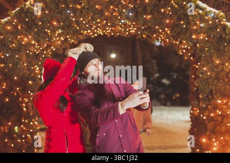 Junge glücklich, Mann und Frau, die selfie mit lustigen Gesichtern in der Nähe von Bogen mit Weihnachten Dekoration, Outdoor im Winter Markt Stockfoto
