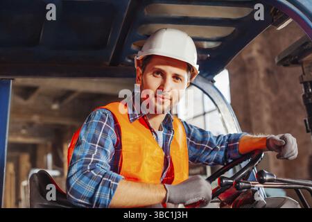 Porträt eines jungen, hübschen Lagerarbeiters, der in einem Froklift-Truck sitzt und die Kamera anschaut. Selbstbewusster Gabelstaplerfahrer bei der Arbeit, mit Hardhut und Stockfoto