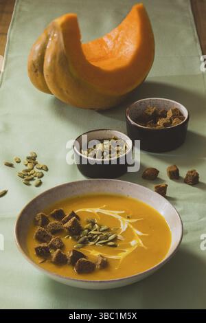 Weiße Keramikschüssel mit Kürbiscreme-Suppe, serviert mit hausgemachten Croutons. Ein Stück Orangenkürbis und zwei Schüsseln mit Kürbiskernen und Croutons Stockfoto