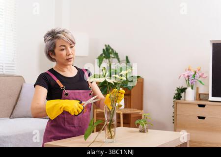Eine ältere asiatische Frau, die eine lila Schürze und gelbe Handschuhe trägt, arbeitet im Innenbereich und schneidet eine Pflanze mit einer Schere. Sie kümmert sich um ihr Haus Stockfoto