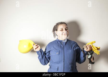 Lächelnde Arbeitnehmerin gekleidet in Arbeitskleidung, hält einen gelben Helm und gelbe Handschuhe Stockfoto
