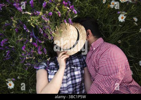 Romantisches Paar, das sich auf dem Sommerfeld mit blühenden Wildblumen umgibt und sich unter dem gewebten Hut küsst Stockfoto