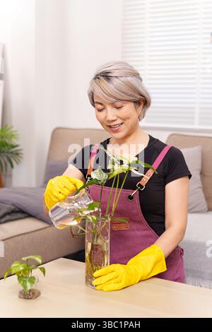 Eine Erwachsene asiatische Frau, die gelbe Handschuhe und eine lila Schürze trägt, lächelt, während sie ihre Zimmerpflanzen wässert und in ihrem Wohnzimmer Pflanzenpflege genießt. Fokus o Stockfoto