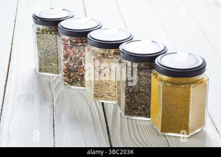 Fünf Glasgefäße mit Gewürzmischungen und getrockneten Kräutern auf einer weißen Holzoberfläche. Auswahl an Gewürzen für Anfänger Stockfoto