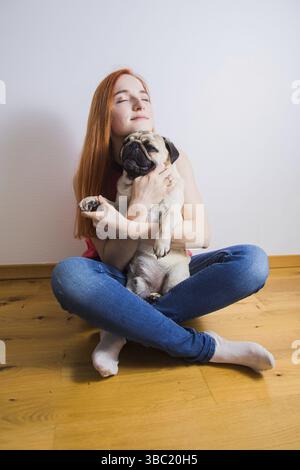Die junge lächelnde rothaarige Frau, die im Zimmer auf dem Boden sitzt und einen Mops hält Stockfoto