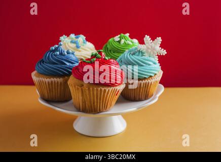 Weißer Kakestand mit fünf Cupcakes mit Sahne bedeckt, auf einem Holztisch. Jeder Cupcake hat seine eigene farbige Glasur oben drauf. Isoliert auf rotem Hintergrund Stockfoto