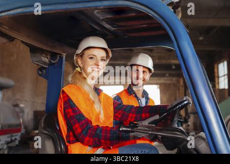 Porträt einer schönen lächelnden Frau, einem Gabelstaplerfahrer, der in einem Lastwagen sitzt und die Kamera anschaut. Junger Mann, Lagerarbeiter, der mit Bewunderung auf ihn blickt Stockfoto