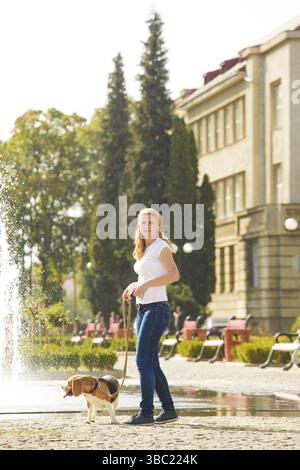 Eine junge Frau, die mit einem Beagle-Hund an der Leine in der Nähe des Brunnens im Park spaziert Stockfoto