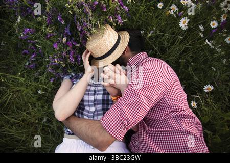 Romantisches Paar, das sich auf dem Sommerfeld mit blühenden Wildblumen umgibt und sich unter dem gewebten Hut küsst Stockfoto