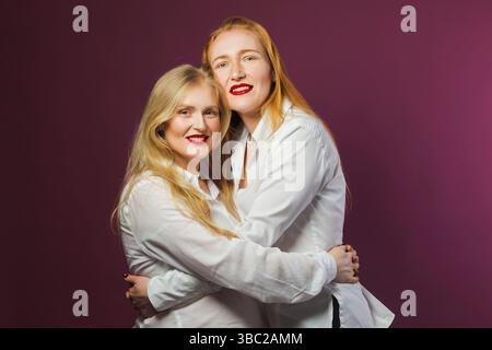 Zwei charmante Frauen, eine junge Mutter und eine Erwachsene Tochter, beide tragen weiße Hemden und roten Lippenstift. Naturschönheitskonzept. Studio mit violettem Ba Stockfoto