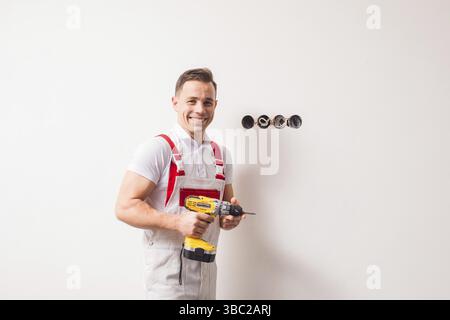 Der starke junge Elektriker hält einen Bohrer. Er steht an den Steckdosen an der weißen Wand, lächelt und schaut in die Kamera Stockfoto