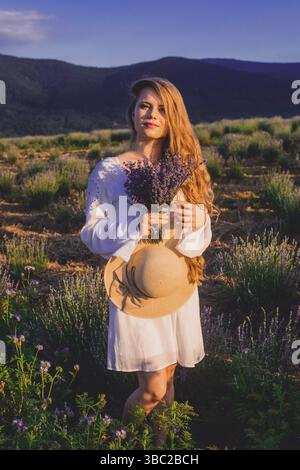 Porträt einer schönen blonden Frau, die zwischen den Plantagen von Lavendel steht. Mädchen in einem weißen Sundress gekleidet und hält sbunch Lavendel Stockfoto