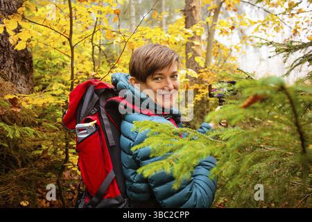 Die lächelnde Wanderer in einer warmen Jacke mit Rucksack macht ein Selfie im Herbstwald Stockfoto