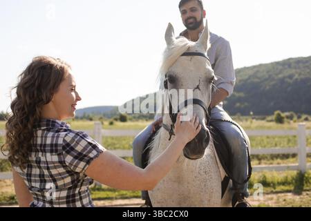 Der gutaussehende bärtige Mann sitzt auf einem Pferd. Und die Frau steht neben einem Pferd und streichelt einen Pferdekopf Stockfoto
