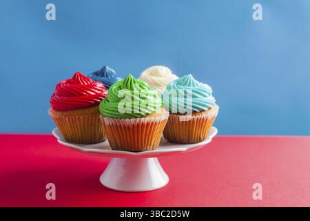 Fünf köstliche Cupcakes mit farbenfroher Glasur auf einem weißen Kakestand, isoliert auf zweifarbigem rot-blauem Hintergrund Stockfoto